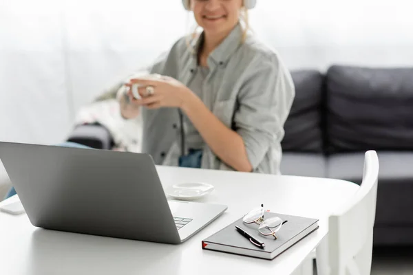 Occhiali e penna in cima al notebook vicino al laptop e allegro freelance sorridente mentre tiene una tazza di caffè su sfondo sfocato a casa, lavoro da casa, spazio di lavoro moderno — Foto stock
