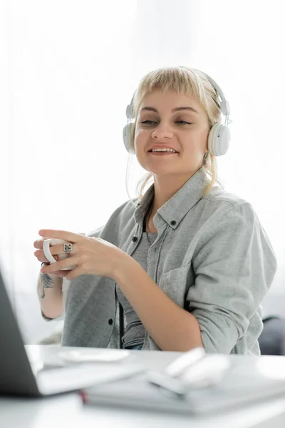 Sonriente mujer joven con el pelo rubio, flequillo y tatuaje en la mano sentado en auriculares inalámbricos y la celebración de la taza de café cerca de la computadora portátil y borrosa portátil y vasos en la mesa, el trabajo desde casa - foto de stock