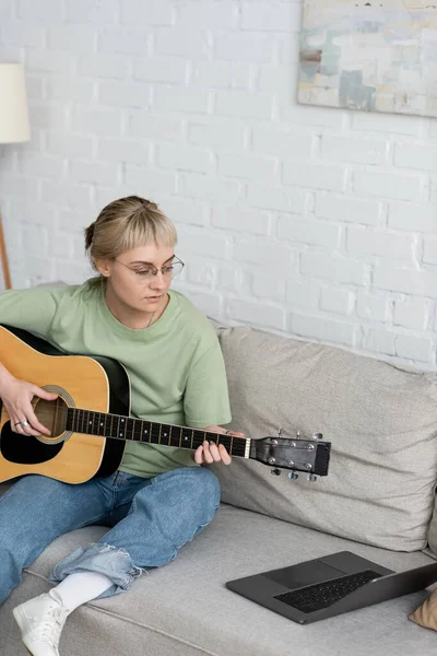 Mujer joven en gafas con flequillo y pelo corto sosteniendo la guitarra acústica y mirando video tutorial en el ordenador portátil y sentado en cómodo sofá en la sala de estar moderna en casa, aprendizaje digital - foto de stock