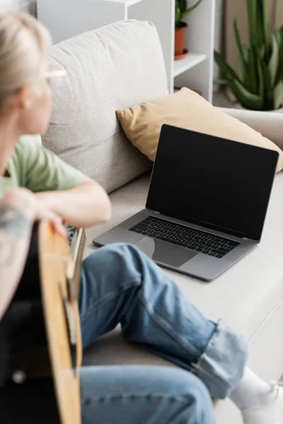 Mujer joven en gafas sosteniendo la guitarra acústica y aprender a tocar mientras busca video tutorial en el ordenador portátil con pantalla en blanco y sentado en un cómodo sofá en la sala de estar, clases de guitarra - foto de stock