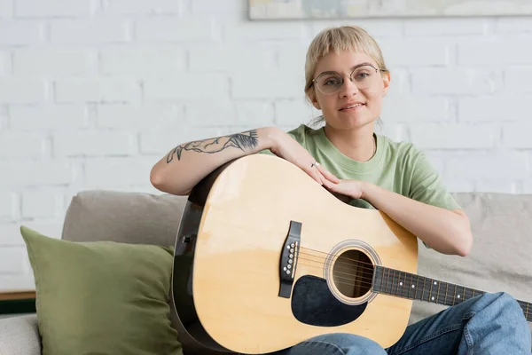 Задоволена молода жінка в окулярах з чубчиком і татуюванням на руці тримає акустичну гітару і дивиться на камеру, сидячи на зручному дивані в сучасній вітальні вдома — стокове фото