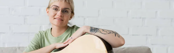 Mujer joven satisfecha en gafas con flequillo y tatuaje en la mano sosteniendo la guitarra acústica y mirando a la cámara mientras está sentado en la sala de estar moderna en casa, sonrisa, pancarta - foto de stock