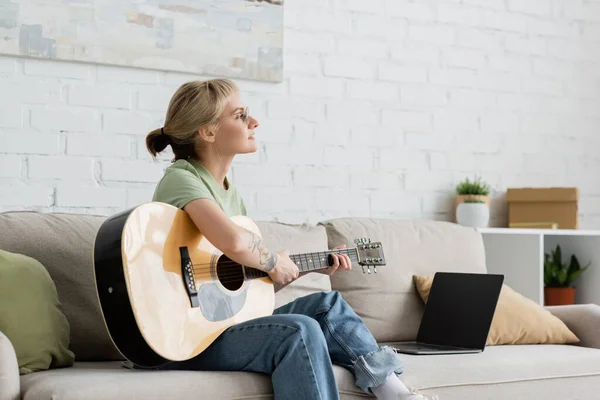 Mujer joven en gafas con flequillo y tatuaje tocando la guitarra acústica cerca de la computadora portátil con pantalla en blanco y sentado en un cómodo sofá en la sala de estar, aprendiendo música, desarrollo de habilidades - foto de stock