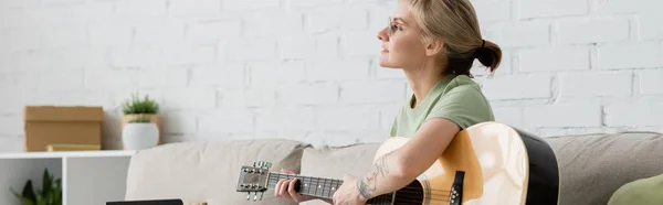 Mujer joven en gafas con flequillo y tatuaje tocando la guitarra acústica y sentado en un cómodo sofá en la sala de estar moderna, aprendiendo música, desarrollo de habilidades, entusiasta de la música, pancarta - foto de stock