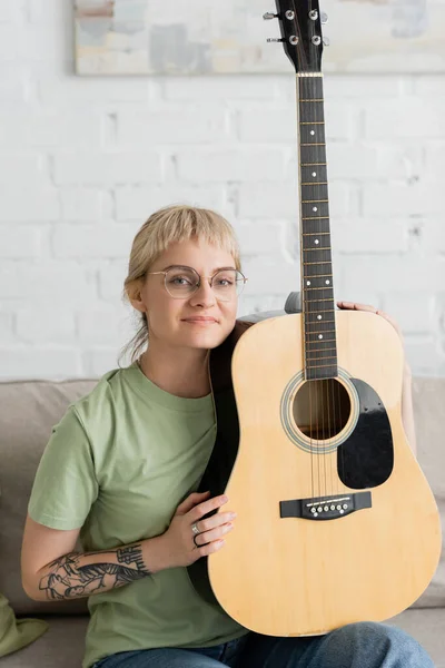 Татуированная и счастливая женщина в очках с челкой, держащая акустическую гитару и сидя на удобном диване в современной гостиной, изучая музыку, развитие навыков, музыкальный энтузиаст, посмотрите на камеру — стоковое фото