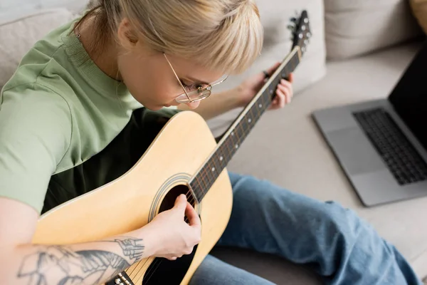 Young woman in glasses with bangs and tattoo holding acoustic guitar and learning how to play near laptop and sitting on comfortable couch in modern living room at home, virtual lessons — Stock Photo