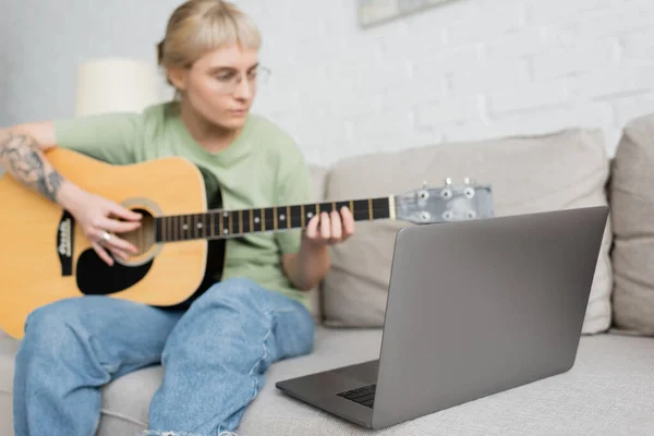 Mujer joven borrosa en gafas con flequillo y tatuaje tocando la guitarra acústica y mirando video tutorial en el ordenador portátil mientras está sentado en un cómodo sofá en la sala de estar moderna, aprendizaje virtual - foto de stock