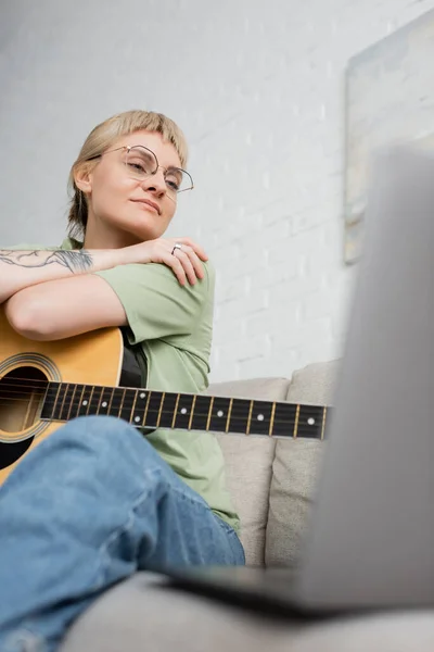 Pleased young woman in glasses with bangs and tattoo holding acoustic guitar and looking video tutorial on laptop while sitting on comfortable couch in modern living room at home, digital resources — Stock Photo