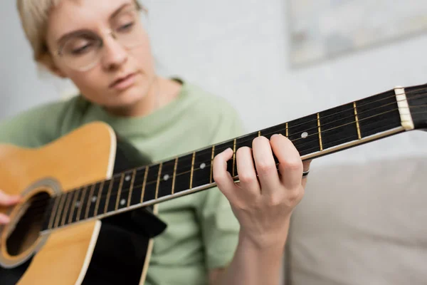 Giovane donna sfocata in occhiali con frangia che suona la chitarra acustica e seduta su un comodo divano in soggiorno moderno, apprendimento della musica, sviluppo delle abilità, appassionato di musica — Foto stock