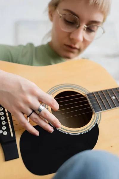 Talanted young woman in glasses with bangs, rings on fingers playing acoustic guitar and sitting in modern living room, learning music, skill development, music enthusiast — Stock Photo