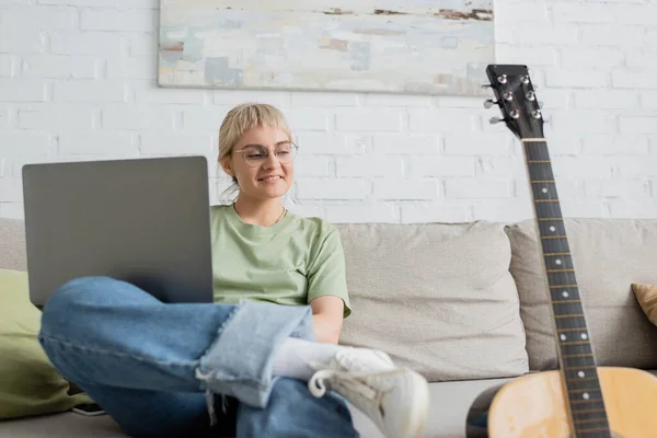 Happy young woman with blonde and short hair, bangs and eyeglasses using laptop while sitting on comfortable couch and looking at guitar in modern living room with painting on wall — Stock Photo