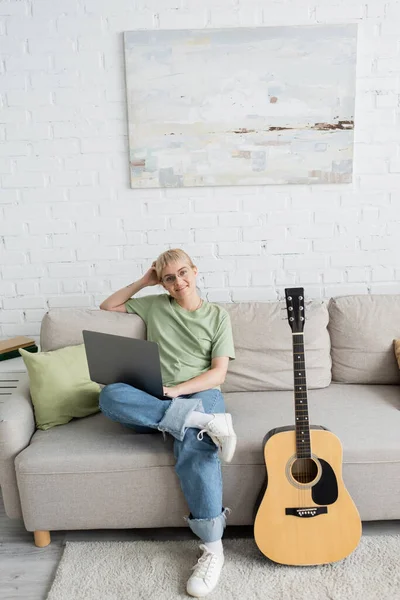 Young woman with blonde and short hair, bangs and eyeglasses using laptop while sitting on comfortable couch and looking at camera near guitar in modern living room with painting on wall — Stock Photo