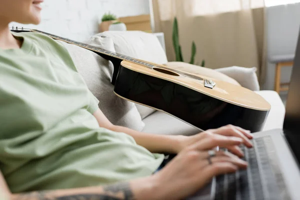 Vista recortada de mujer joven borrosa con tatuaje en la mano usando el ordenador portátil mientras está sentado en un cómodo sofá al lado de la guitarra en la sala de estar moderna, freelance, trabajo desde casa - foto de stock
