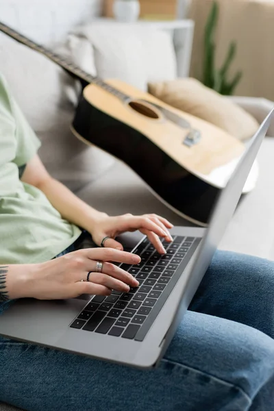 Cropped view of blurred young freelancer with tattoo on hand using laptop while sitting on comfortable couch next to guitar in modern living room, freelance, work from home — Stock Photo