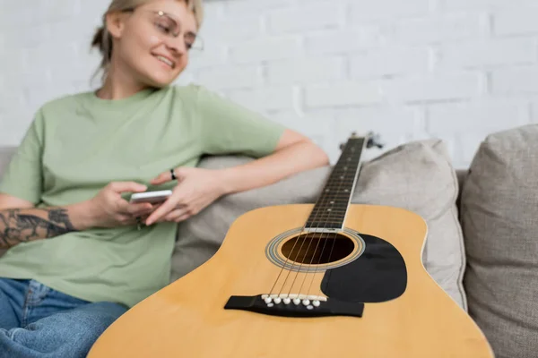 Happy young woman with blonde and short hair, bangs and eyeglasses using smartphone while sitting on comfortable couch near guitar in modern living room, blurred shot — Stock Photo