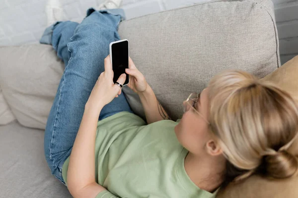 Mujer joven con cabello rubio y corto y anteojos, tatuaje en la mano y ropa casual usando teléfono inteligente mientras descansa en un cómodo sofá, vaqueros, camiseta - foto de stock
