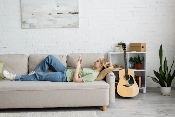 Side view of happy young woman with blonde and short hair, bangs and eyeglasses using smartphone while resting on comfortable couch near guitar in modern living room with painting on wall and plants — Stock Photo