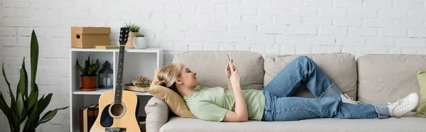 Side view of happy woman with blonde and short hair, bangs and eyeglasses using smartphone while resting on comfortable couch near guitar in modern living room with rack and plants, banner — Stock Photo