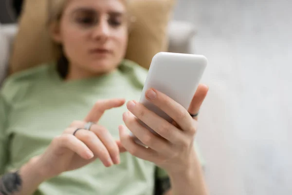 Overhead view of young woman with tattoo, blonde and short hair, bangs and eyeglasses using smartphone while resting on comfortable couch in modern living room, blurred background — Stock Photo
