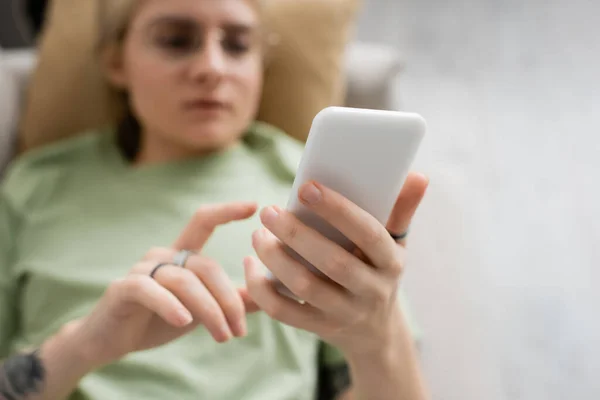 Vue aérienne de la jeune femme avec tatouage, cheveux blonds et courts, frange et lunettes en utilisant smartphone tout en se reposant sur un canapé confortable dans le salon moderne, fond flou — Photo de stock