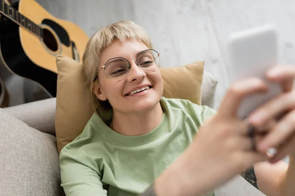 Над головой вид счастливой молодой женщины с светлыми и короткими волосами, челкой и очками смс на смартфоне, отдыхающей на удобном диване рядом с гитарой в современной гостиной — стоковое фото