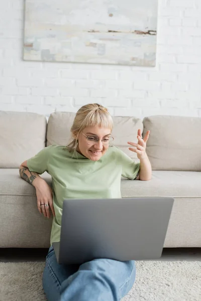 Femme tatouée avec des cheveux blonds et courts, frange et lunettes souriant pendant appel vidéo sur ordinateur portable tout en étant assis sur le tapis près du canapé confortable dans le salon moderne avec paiting sur le mur — Photo de stock
