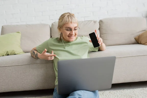 Весела і татуйована жінка з чубчиком і окулярами, використовуючи ноутбук, сидячи на килимі і тримаючи смартфон з порожнім екраном біля зручного дивана в сучасній вітальні — стокове фото