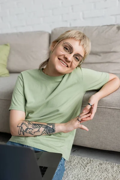 Mujer tatuada tappy con cabello rubio y corto, flequillo y gafas usando el ordenador portátil mientras está sentado en la alfombra cerca de cómodo sofá en la sala de estar moderna y mirando a la cámara - foto de stock