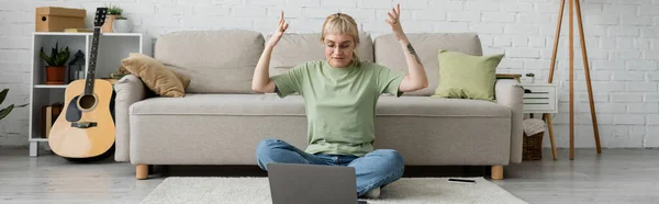 Mujer tatuada emocional con flequillo y gafas usando el ordenador portátil mientras gesticulaba, sentado en la alfombra cerca del teléfono inteligente, cómodo sofá, guitarra y estante con plantas en la sala de estar moderna, pancarta - foto de stock