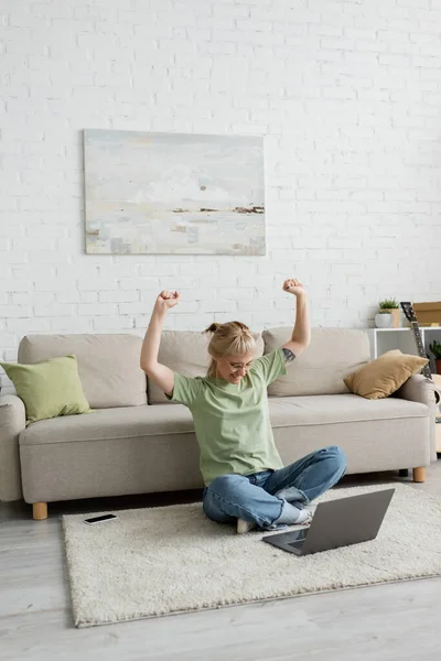 Excited tattooed woman in eyeglasses using laptop while sitting with raised hands on carpet near smartphone, comfortable couch, guitar and rack with plants in modern living room with paiting on wall — Stock Photo
