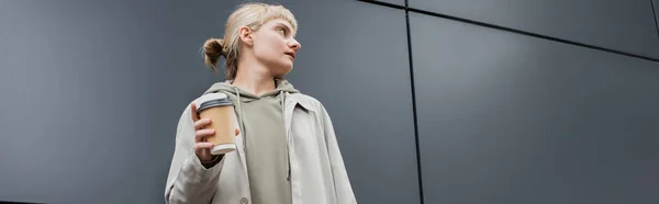 Jeune femme à la mode avec les cheveux blonds avec frange debout dans le manteau et sweat à capuche tout en tenant tasse en papier avec café à emporter près du bâtiment moderne gris dans la rue, à l'extérieur, vie urbaine, bannière — Photo de stock