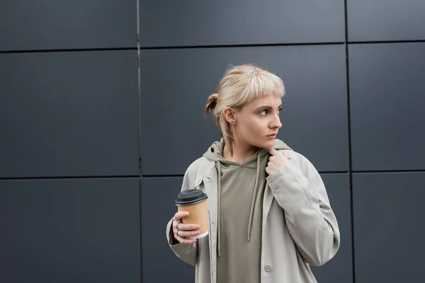 Mujer joven de moda con cabello rubio con flequillo de pie en abrigo y sudadera con capucha mientras sostiene la taza de papel con café para llevar cerca de edificio moderno gris en la calle, exterior, vida urbana - foto de stock