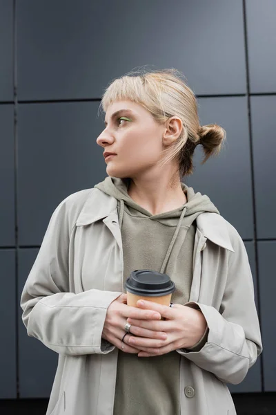 Stylish young woman with blonde hair with bangs standing in coat and hoodie while holding paper cup with takeaway coffee near grey modern building on street, outside, urban living, look away — Stock Photo