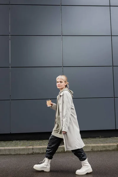 Full length of trendy woman with blonde hair with bangs walking in coat, black leather pants, hoodie and boots while holding paper cup with takeaway coffee near grey modern building on street — Stock Photo