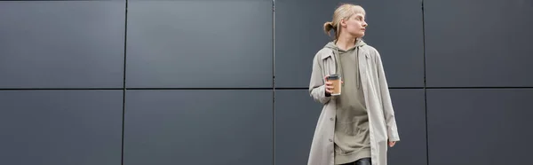 Stylische junge Frau mit Pony in Mantel und Kapuzenpulli, während sie Pappbecher mit Kaffee zum Mitnehmen in der Nähe eines grauen modernen Gebäudes auf der Straße hält, draußen, urbanes Leben, wegschauen, Transparent — Stockfoto