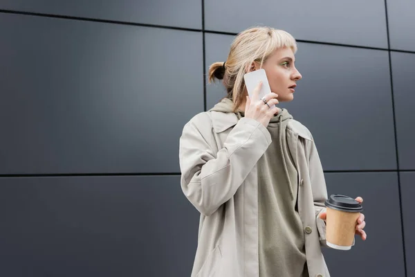 Модная молодая женщина с челкой и светлые волосы держа бумажный стаканчик с кофе, чтобы пойти во время разговора на смартфоне и стоя в толстовке и пальто возле серого современного здания на городской улице, отвернуться — стоковое фото