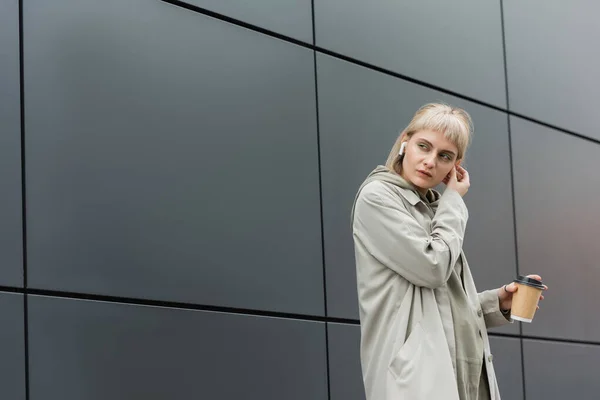 Blonde young woman with bangs holding paper cup with coffee to go while adjusting wireless earphones and standing in trendy outfit, hoodie and coat near grey modern building on urban street — Stock Photo