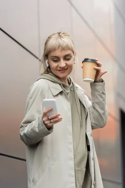 Happy young woman with bangs holding paper cup with coffee to go and standing in trendy outfit and wireless earphones while using smartphone near grey modern building on urban street — Stock Photo