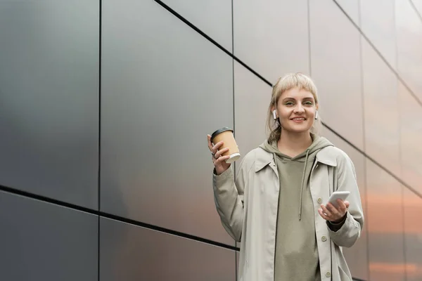 Усміхнена молода жінка з чубчиком тримає паперову чашку з кавою, щоб піти і стояти в модному вбранні та бездротових навушниках, використовуючи смартфон біля сірої сучасної будівлі на міській вулиці, подивіться на камеру — стокове фото