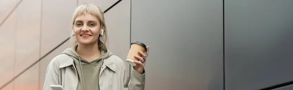 Sonriente mujer joven con flequillo sosteniendo taza de papel con café para ir y de pie en traje de moda y auriculares inalámbricos mientras se utiliza el teléfono inteligente cerca de edificio moderno gris en la calle urbana, pancarta - foto de stock