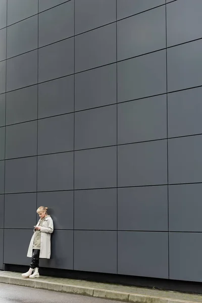 Молода жінка з чубчиком стоїть в модному одязі та бездротових навушниках, використовуючи смартфон біля сірої сучасної будівлі на міській вулиці, пальто з светром, чорні шкіряні штани та чоботи, повна довжина — стокове фото