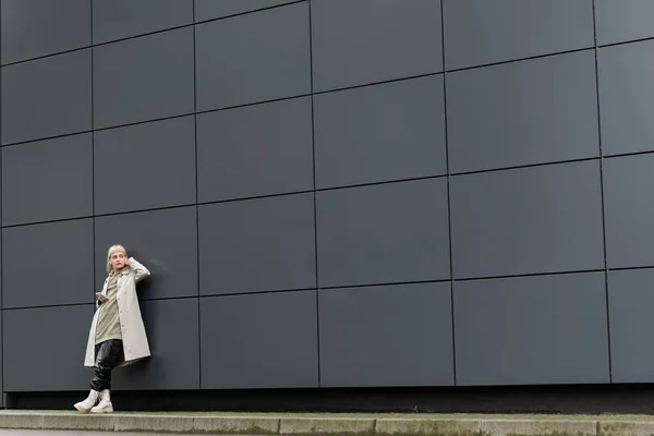 Молодая женщина с челкой, стоящая в стильной одежде и беспроводных наушниках, используя смартфон возле серого современного здания на городской улице, пальто с толстовкой, черные кожаные брюки и сапоги, полная длина — стоковое фото