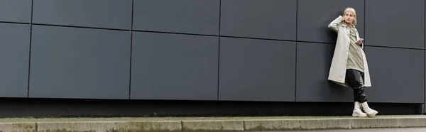 Junge Frau mit Pony in trendiger Kleidung und kabellosen Kopfhörern, während sie das Smartphone in der Nähe des grauen modernen Gebäudes an der Stadtstraße benutzt, Mantel mit Kapuzenpulli, schwarze Lederhose und Stiefel, Banner — Stockfoto