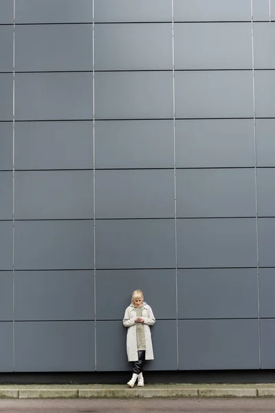 Блондинка с челкой, стоящая в модной одежде и беспроводных наушниках, используя смартфон возле серого современного здания на городской улице, пальто с толстовкой, черные кожаные брюки и сапоги, полная длина — стоковое фото