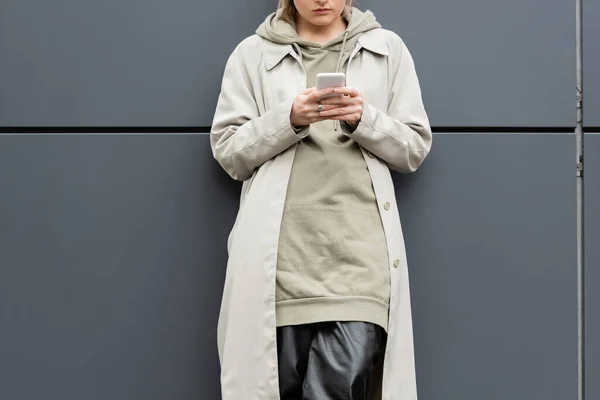 Обрізаний вид модної молодої жінки, що стоїть в модному одязі та смс на смартфоні біля сірої стіни сучасної будівлі на міській вулиці, пальто з капюшоном, частковий вид — стокове фото