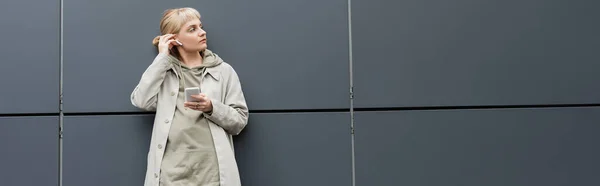 Elegante giovane donna con frangia in piedi in abiti alla moda e auricolari wireless durante l'utilizzo di smartphone vicino grigio edificio moderno sulla strada urbana, cappotto con cappuccio, guardando altrove, banner — Foto stock