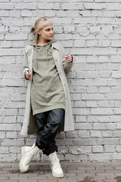 Fashonable young woman with makeup, blonde hair, bangs, in stylish outfit, long hoodie, coat, black leather pants and beige boots standing near grey brick wall and looking away — Stock Photo