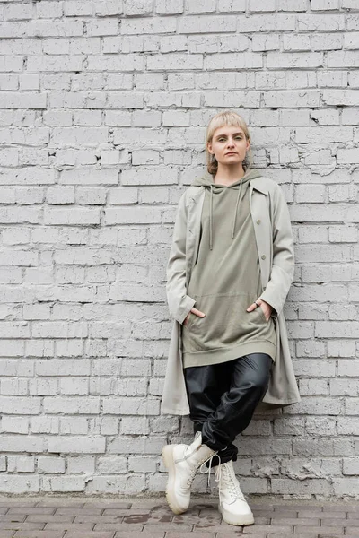 Young woman with makeup, blonde hair, bangs, in stylish outfit, long hoodie, coat, black leather pants and beige boots standing with hands in pockets near grey brick wall and looking at camera — Stock Photo
