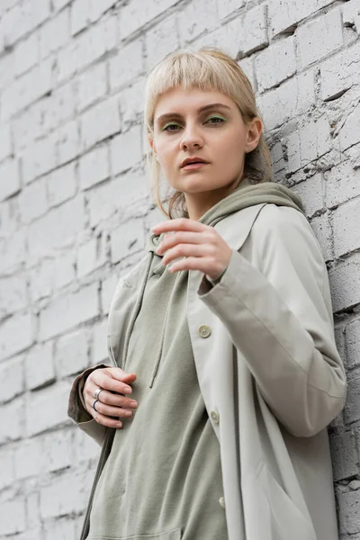 Young woman with makeup, blonde hair, bangs, in stylish outfit standing near grey brick wall of modern building and looking at camera while posing outdoor, coat, fashion trend — Stock Photo