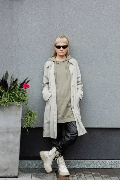 Full length of young woman in stylish sunglasses, long hoodie, coat, black leather pants and beige boots standing near grey wall of modern building and posing with hands in pockets near green plant — Stock Photo